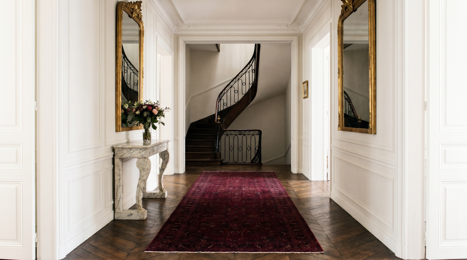 Couloir haussmannien parisien avec long tapis sur-mesure bordeaux 80x800 cm sur parquet ancien chevron, rampe escalier fer forgé et console marbre