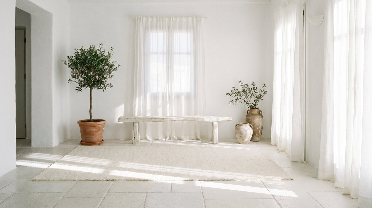 Intérieur méditerranéen Cyclades avec sol pierre blanche calcaire, tapis blanc ivoire 200x300, banc bois flotté, plante olivier en pot terre cuite et mur en chaux blanche