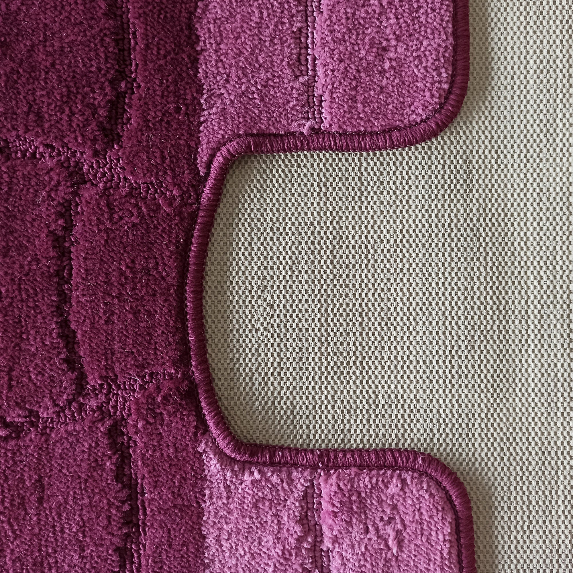 Tapis aux teintes magenta et pourpre à texture bouclée et motifs en relief, avec un bord arrondi sur un fond beige à motif quadrillé