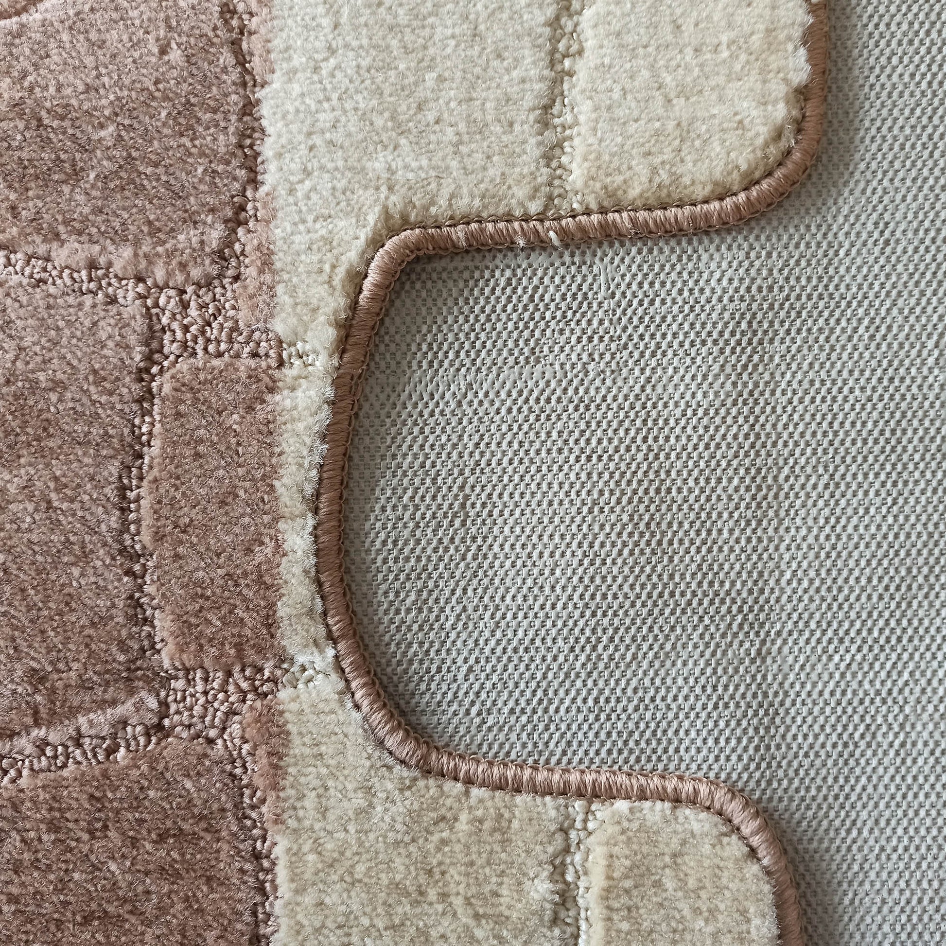 Tapis texturé à poils longs alternant des zones crème et marron clair. Une bordure rose ondulée sépare cette surface d'un motif quadrillé