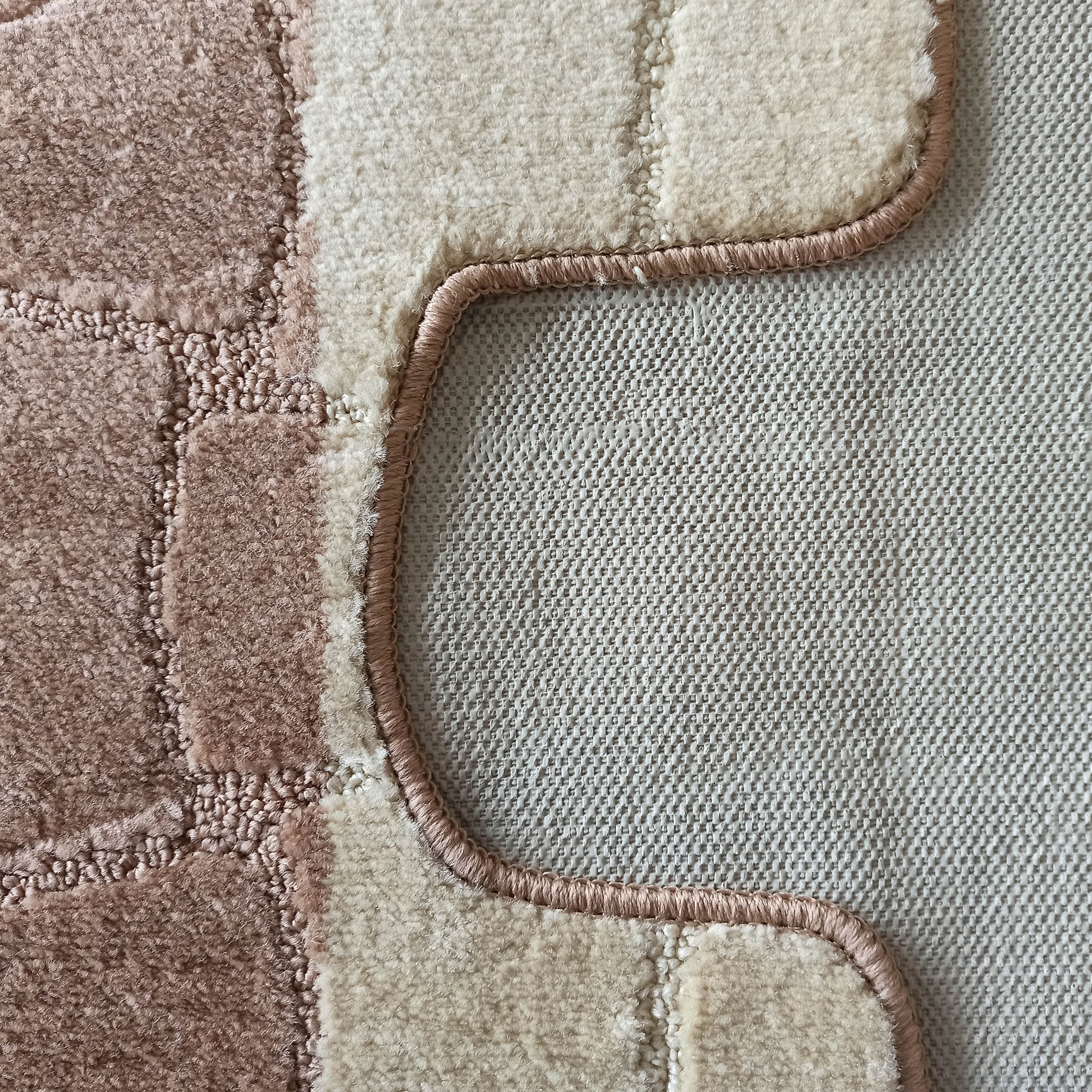 Tapis texturé à poils longs alternant des zones crème et marron clair. Une bordure rose ondulée sépare cette surface d'un motif quadrillé