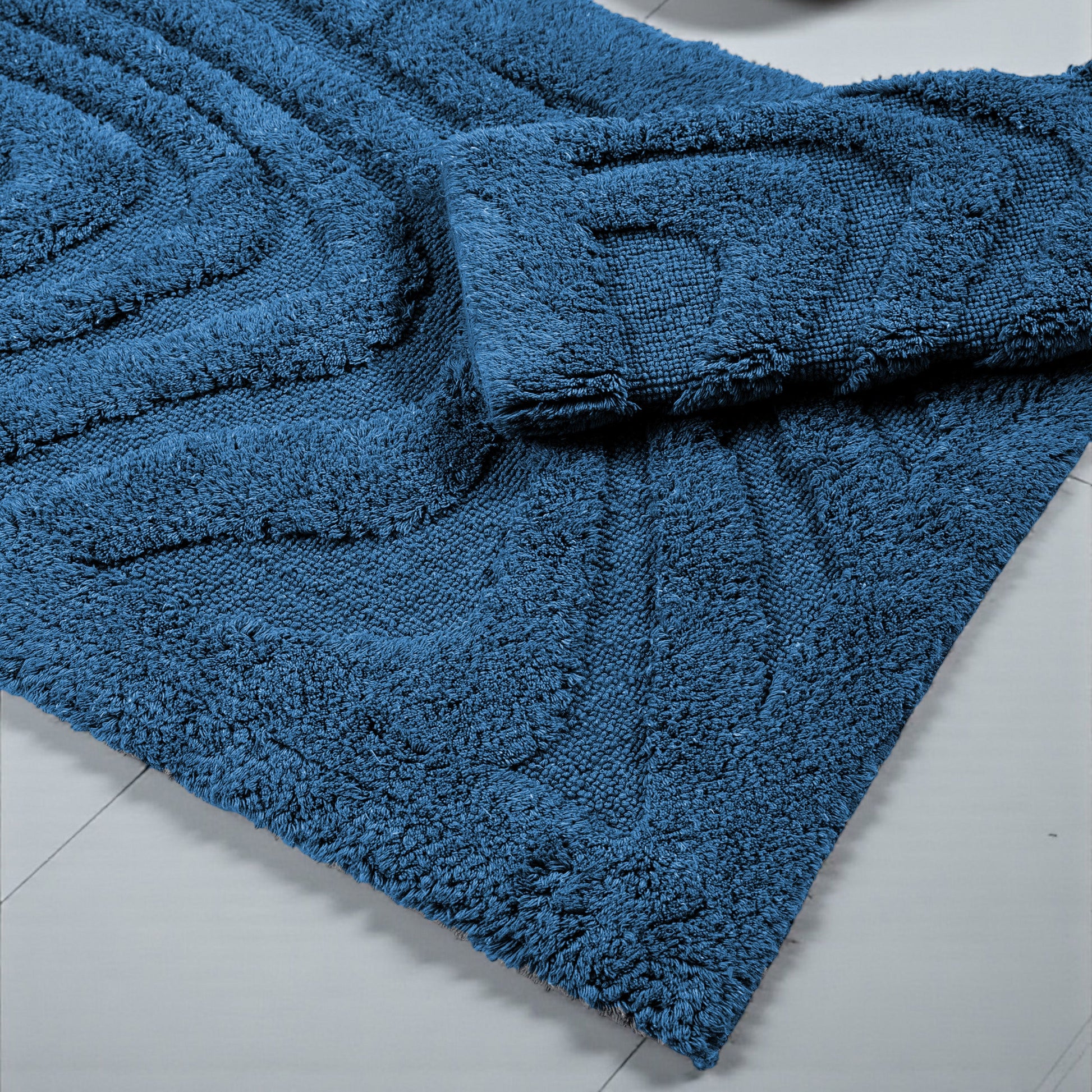 Tapis bleu profond, texture douce et bouclée, motifs en relief de vagues diagonales, partiellement replié sur carrelage clair