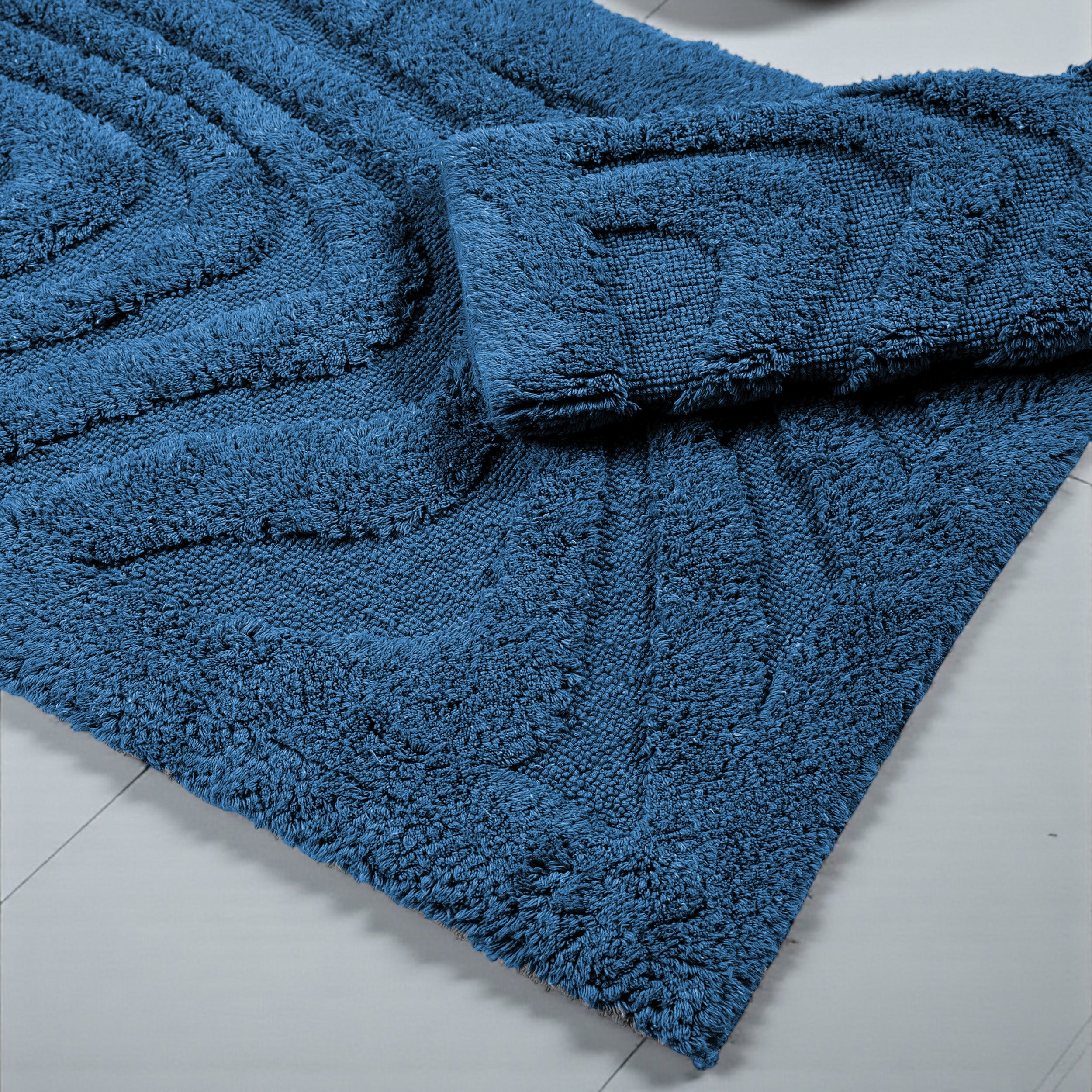 Tapis bleu profond, texture douce et bouclée, motifs en relief de vagues diagonales, partiellement replié sur carrelage clair