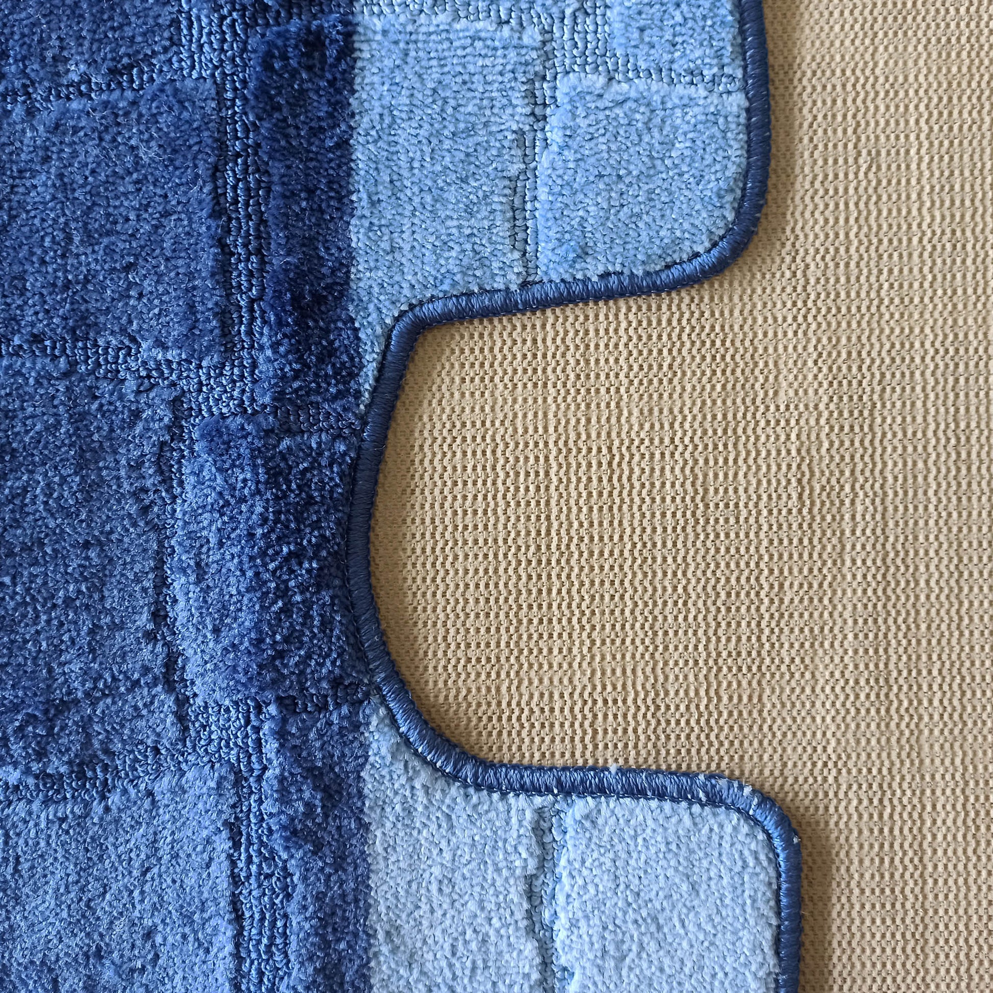 Tapis bleu texturé aux nuances claires et foncées, avec des carrés en relief et une bordure ondulée sur un fond beige tissé