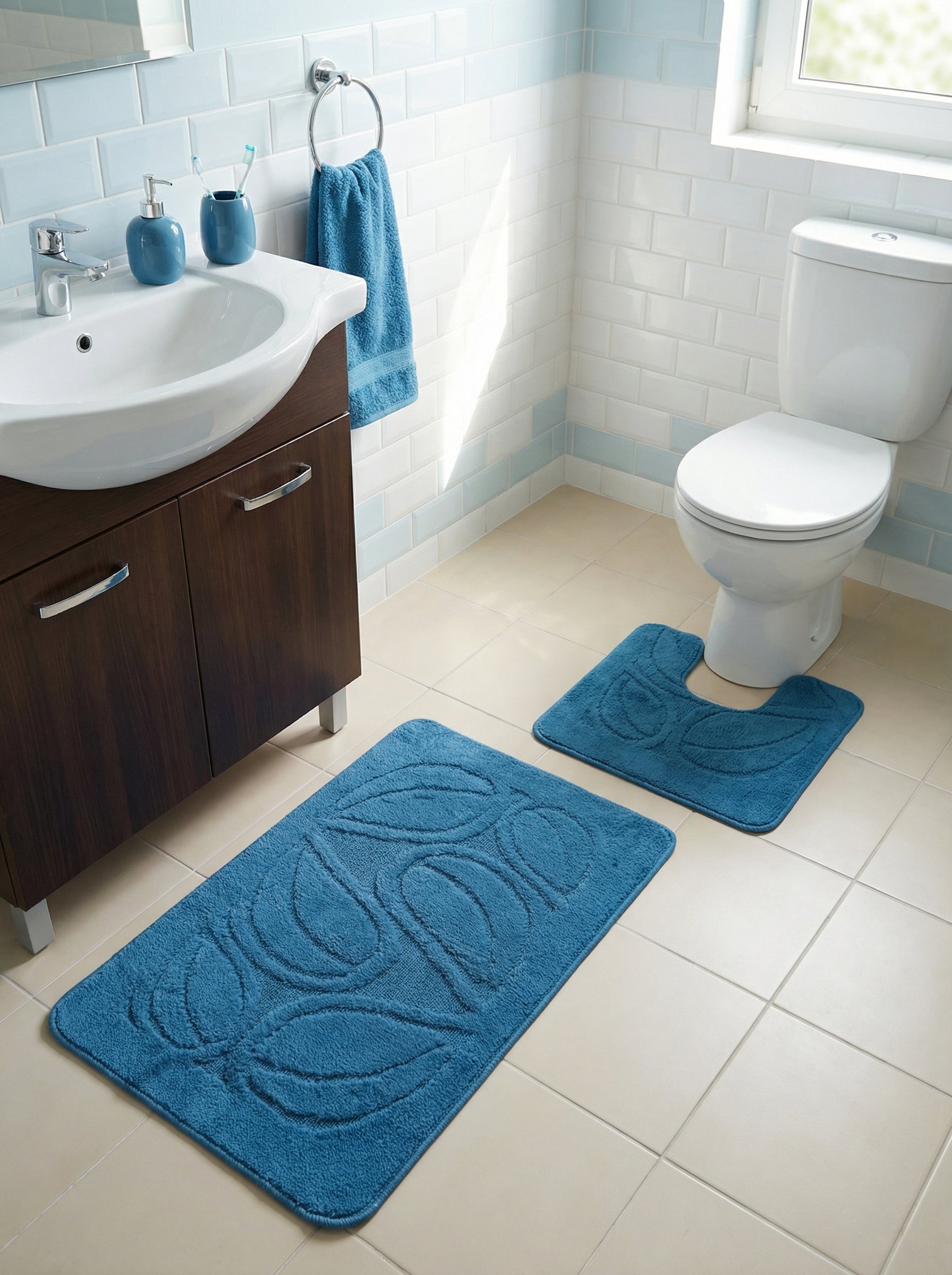Deux tapis de bain bleus en tissu éponge avec motifs de feuilles, un rectangulaire et un en U, sur le sol carrelé d'une salle de bain