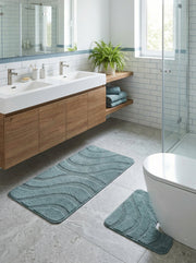 Deux tapis de bain bleu sarcelle à motifs ondulés et texturés, un rectangulaire et un en U, posés sur un sol carrelé gris dans une salle de bain moderne