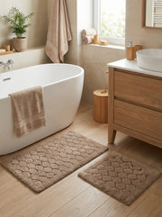 Deux tapis de bain beiges, un grand et un petit, avec des motifs de galets en relief, posés sur un plancher en bois clair dans une salle de bain
