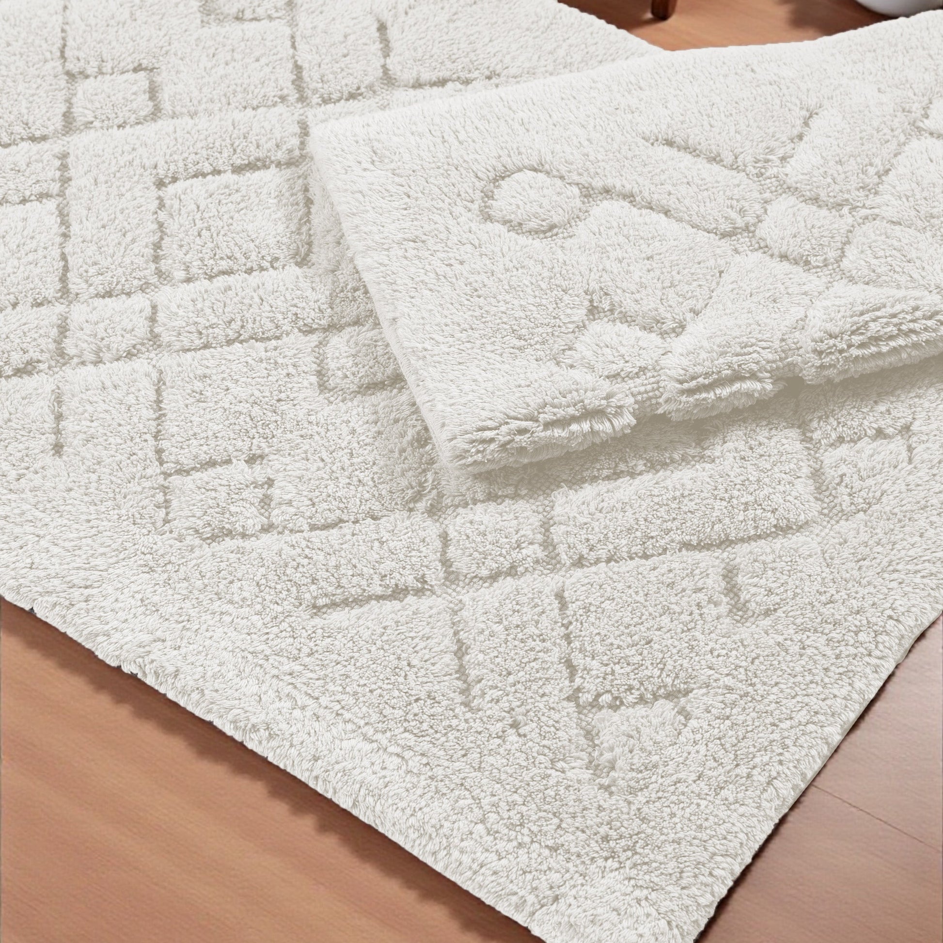 Tapis blanc cassé moelleux aux motifs géométriques en relief, partiellement plié sur un plancher en bois