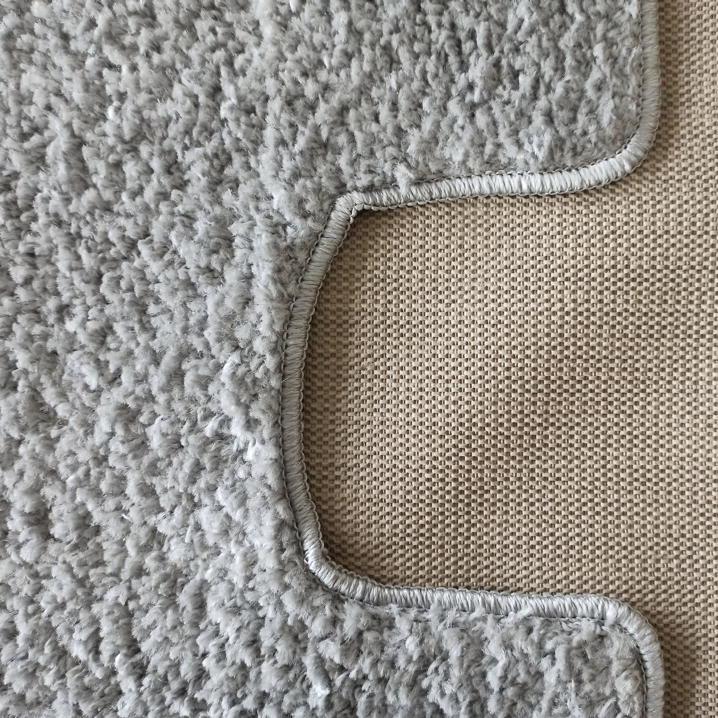 Gros plan d'un tapis gris clair à poils longs, avec un bord surpiqué visible sur un fond beige tissé