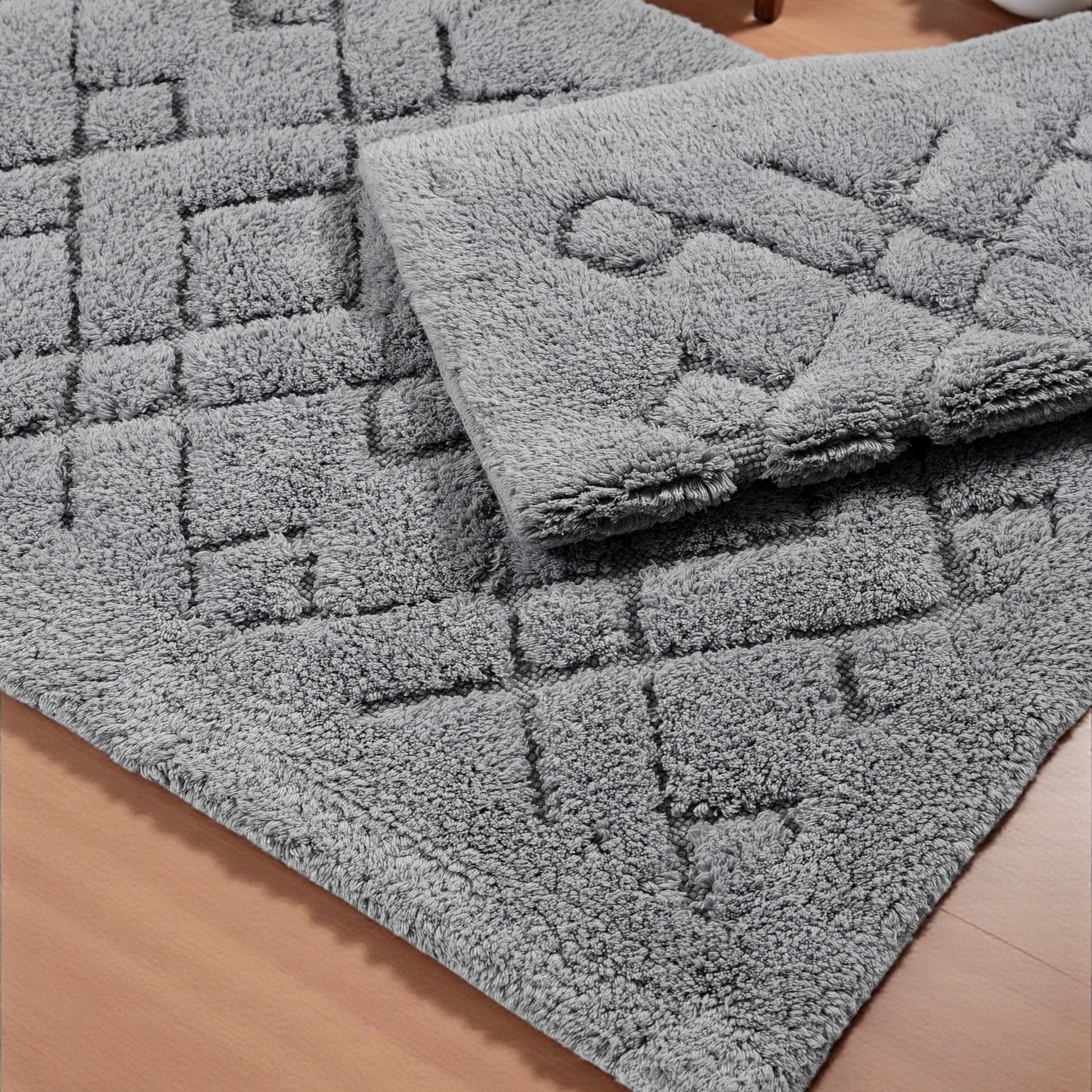 Deux tapis gris moelleux, l'un avec un motif de carrés en relief, l'autre plié, posés sur un plancher en bois