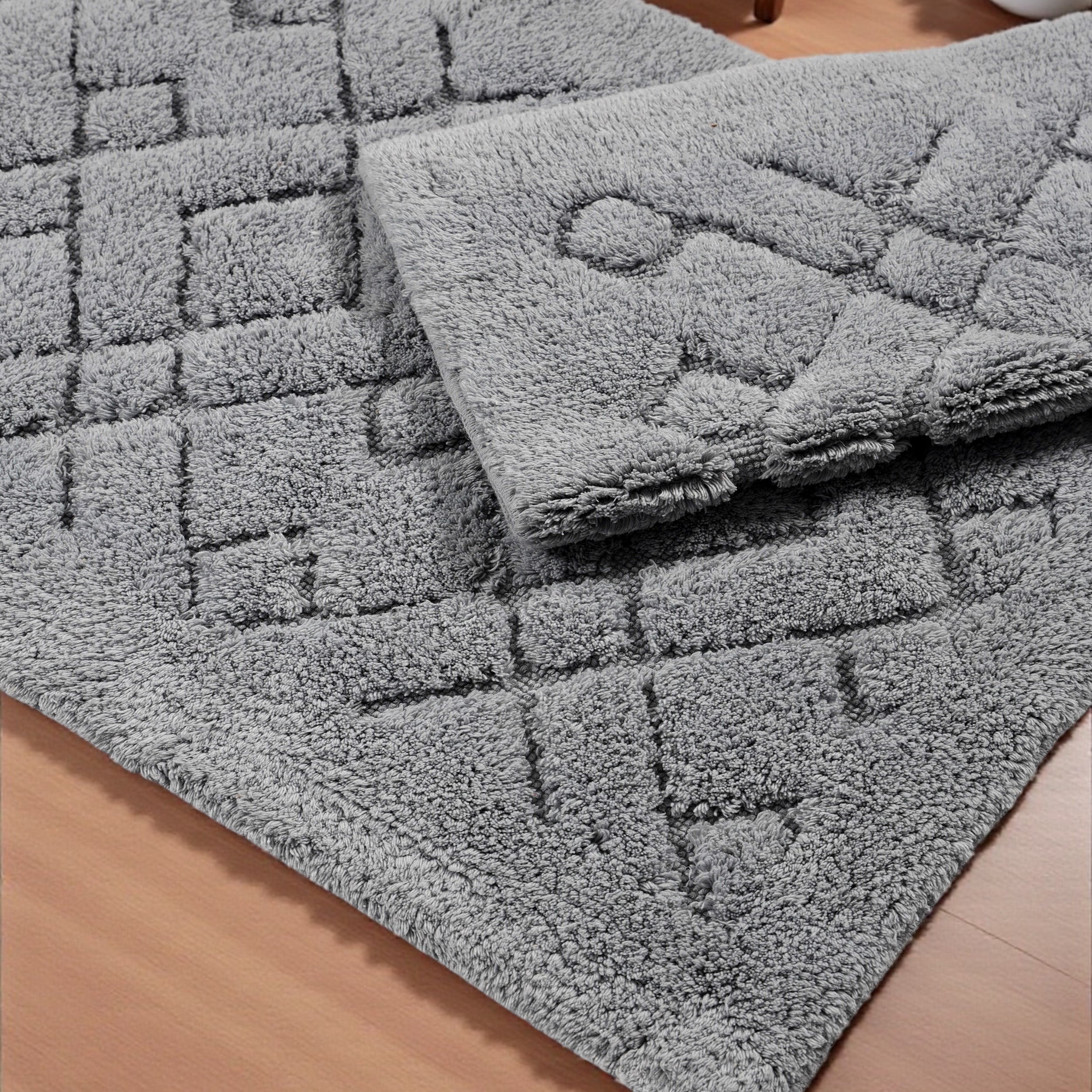 Deux tapis gris moelleux, l'un avec un motif de carrés en relief, l'autre plié, posés sur un plancher en bois
