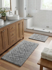 Deux tapis de bain gris rectangulaires à motifs géométriques en relief sur un sol en bois clair dans une salle de bain