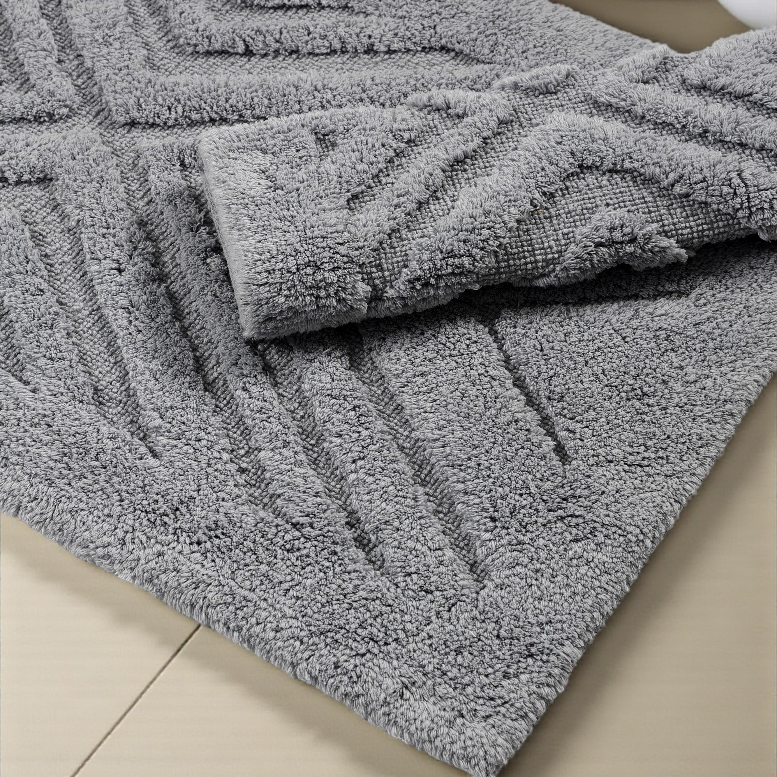Tapis gris moelleux à texture bouclée, avec un motif de chevrons en relief, une partie est repliée sur un sol clair