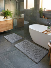 Deux tapis de bain rectangulaires gris avec des motifs texturés de galets, posés sur un sol carrelé foncé dans une salle de bain