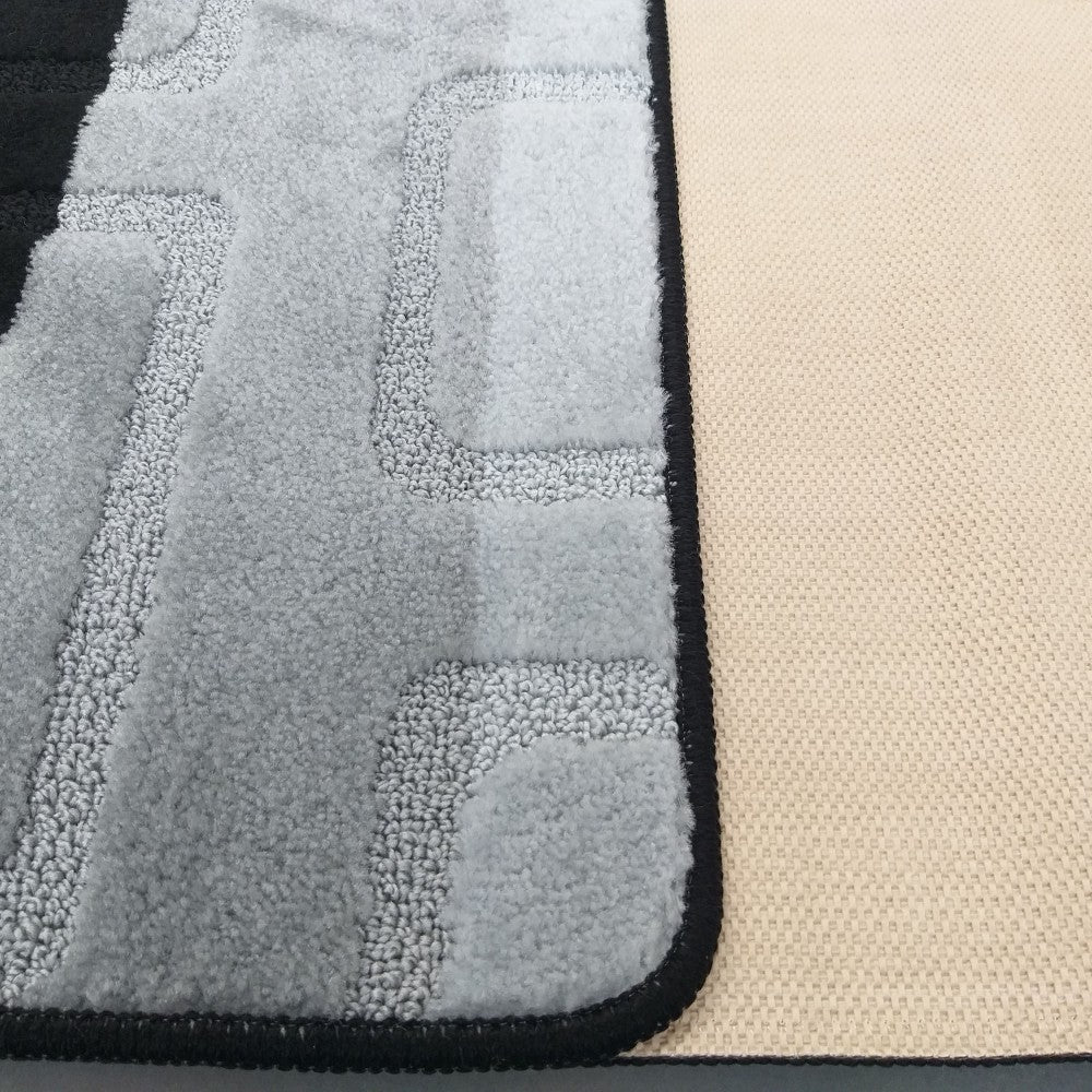 Tapis gris doux avec des motifs géométriques en relief, bordé de noir. L'envers tissé beige est visible à droite