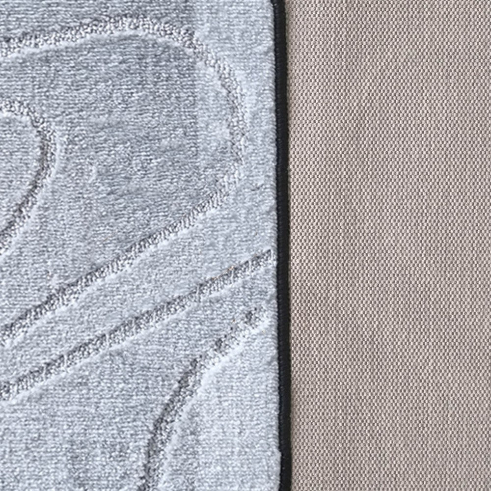 Tapis gris clair texturé avec des motifs sculptés, bordé de noir. Le revers du tapis, visible à droite, est beige et finement tissé