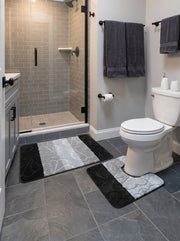 Ensemble de deux tapis de bain moelleux, un rectangulaire et un contour de toilettes. Ils sont noirs et gris clair, avec des motifs floraux en relief. Ils sont posés sur le carrelage foncé d'une salle de bain