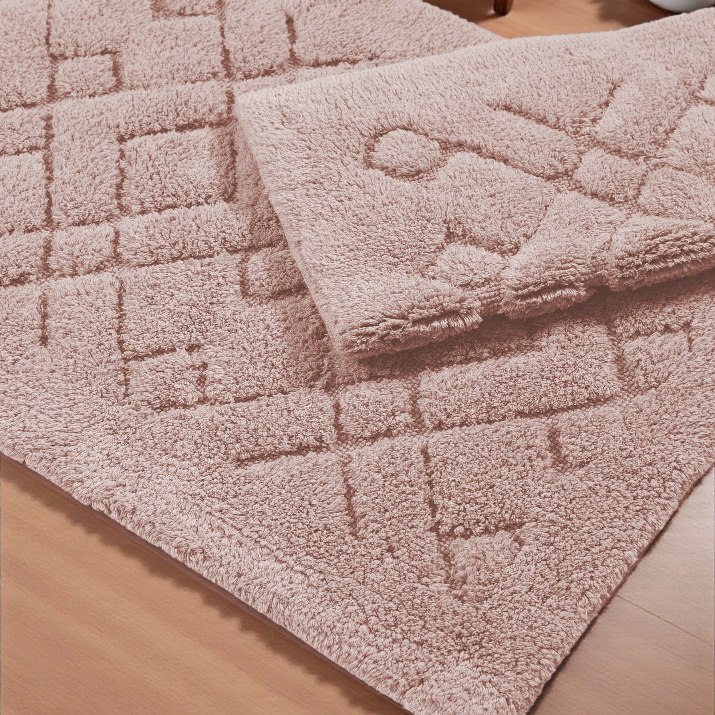 Deux tapis roses en textile doux, l'un à motifs carrés, l'autre plié à motifs circulaires en relief sur un plancher de bois