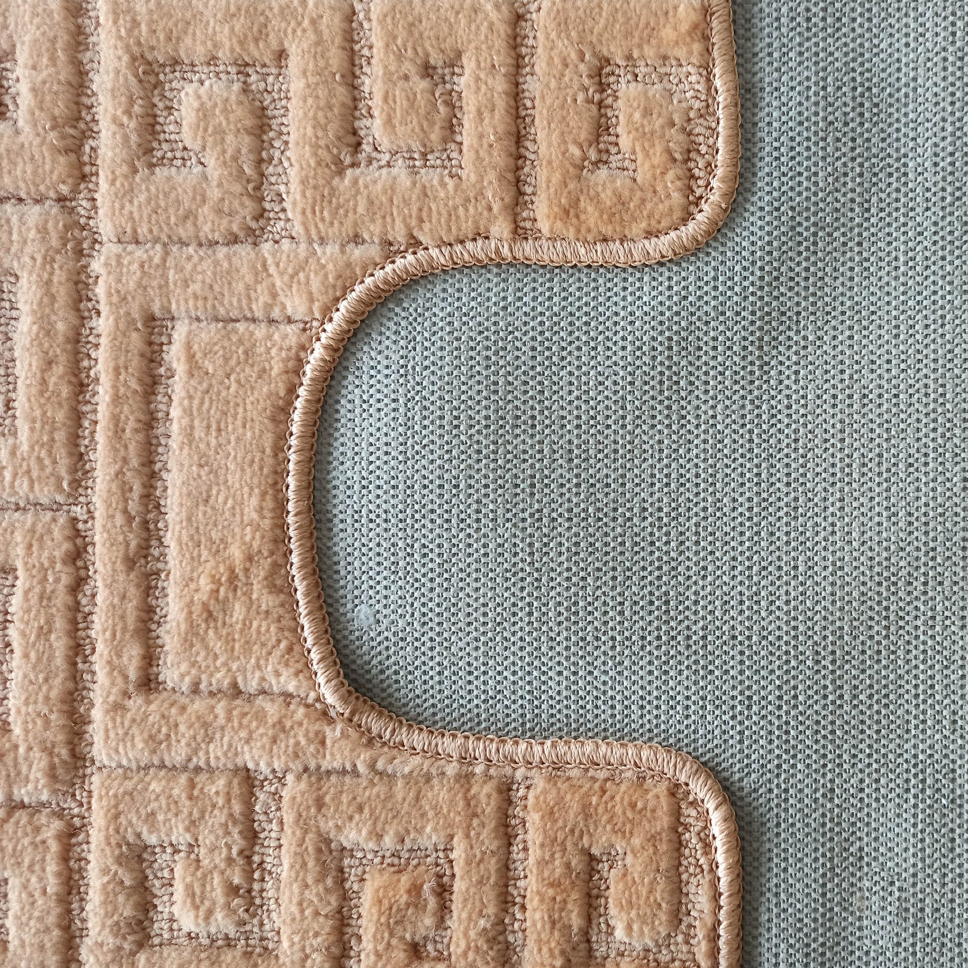 Tapis beige clair à texture douce avec motifs de méandres en relief, bordé d'un liseré incurvé sur son dos gris texturé