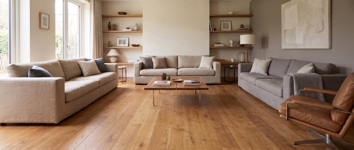 Vaste salon lumineux aux murs crème et gris, avec parquet clair, canapés beiges et gris, étagères murales, et sol nu