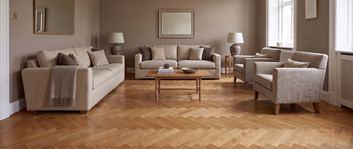 Dans un salon aux teintes douces, le sol est un parquet à chevrons sous des canapés et fauteuils clairs, sans tapis