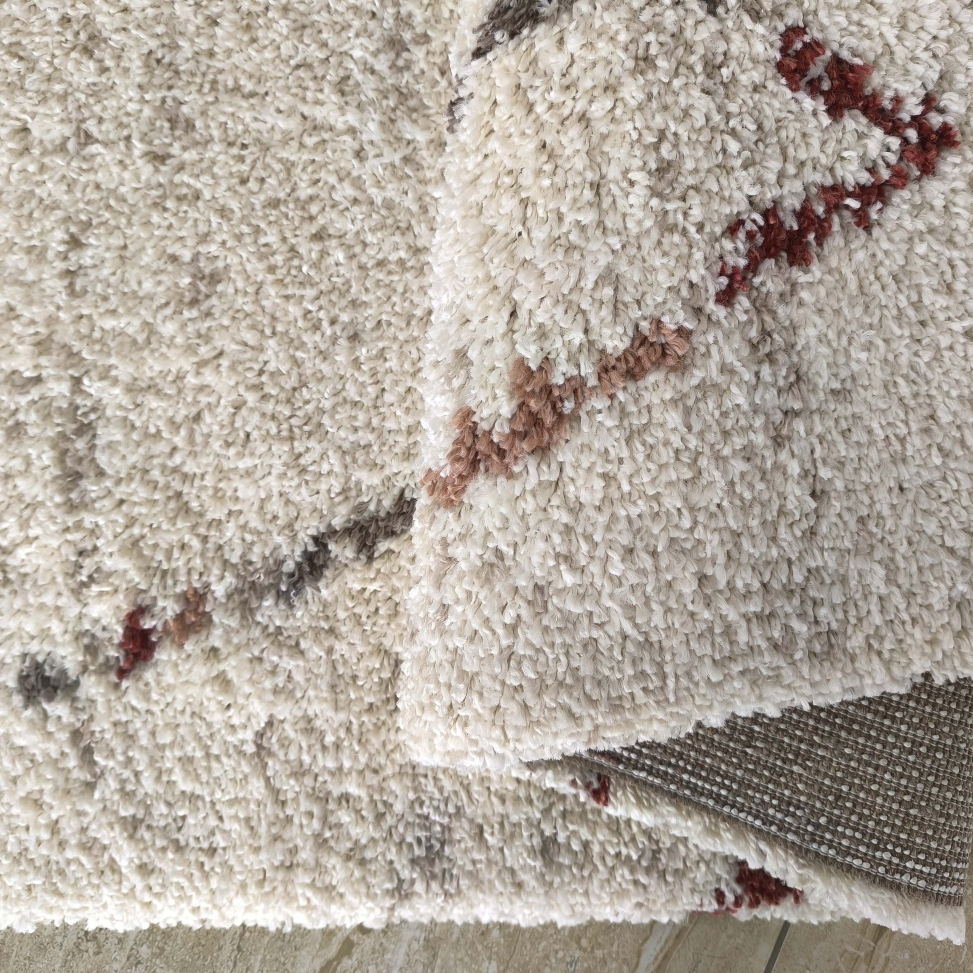 Tapis à poils longs couleur crème avec des motifs abstraits en marron et rouge brique, une partie est pliée laissant voir son dos tissé