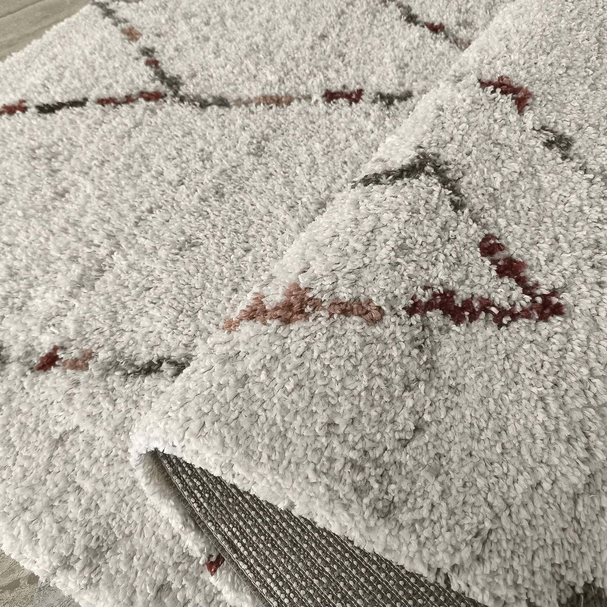 Tapis shaggy gris clair orné de motifs à lignes brisées en gris foncé, bordeaux et brun rosé, une partie est repliée