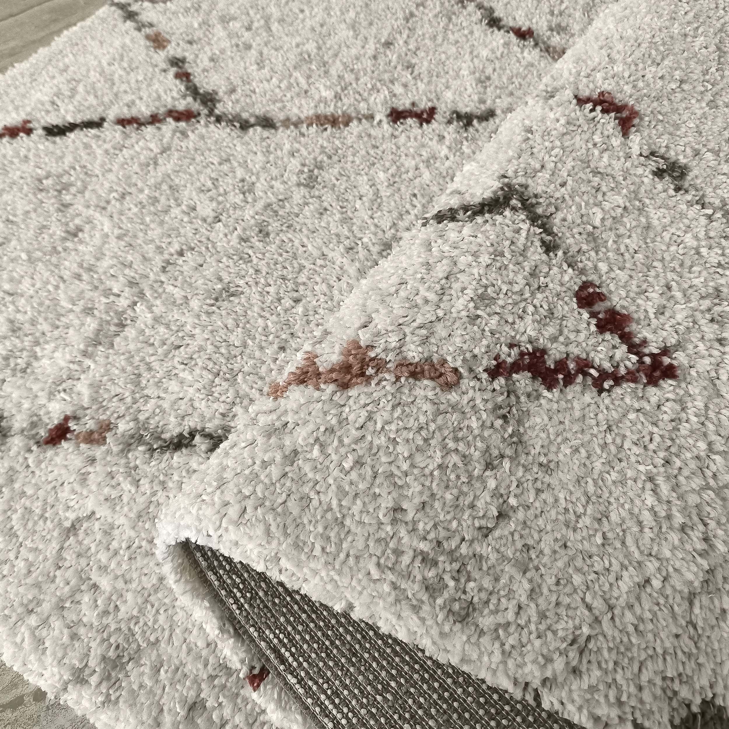Tapis shaggy gris clair orné de motifs à lignes brisées en gris foncé, bordeaux et brun rosé, une partie est repliée