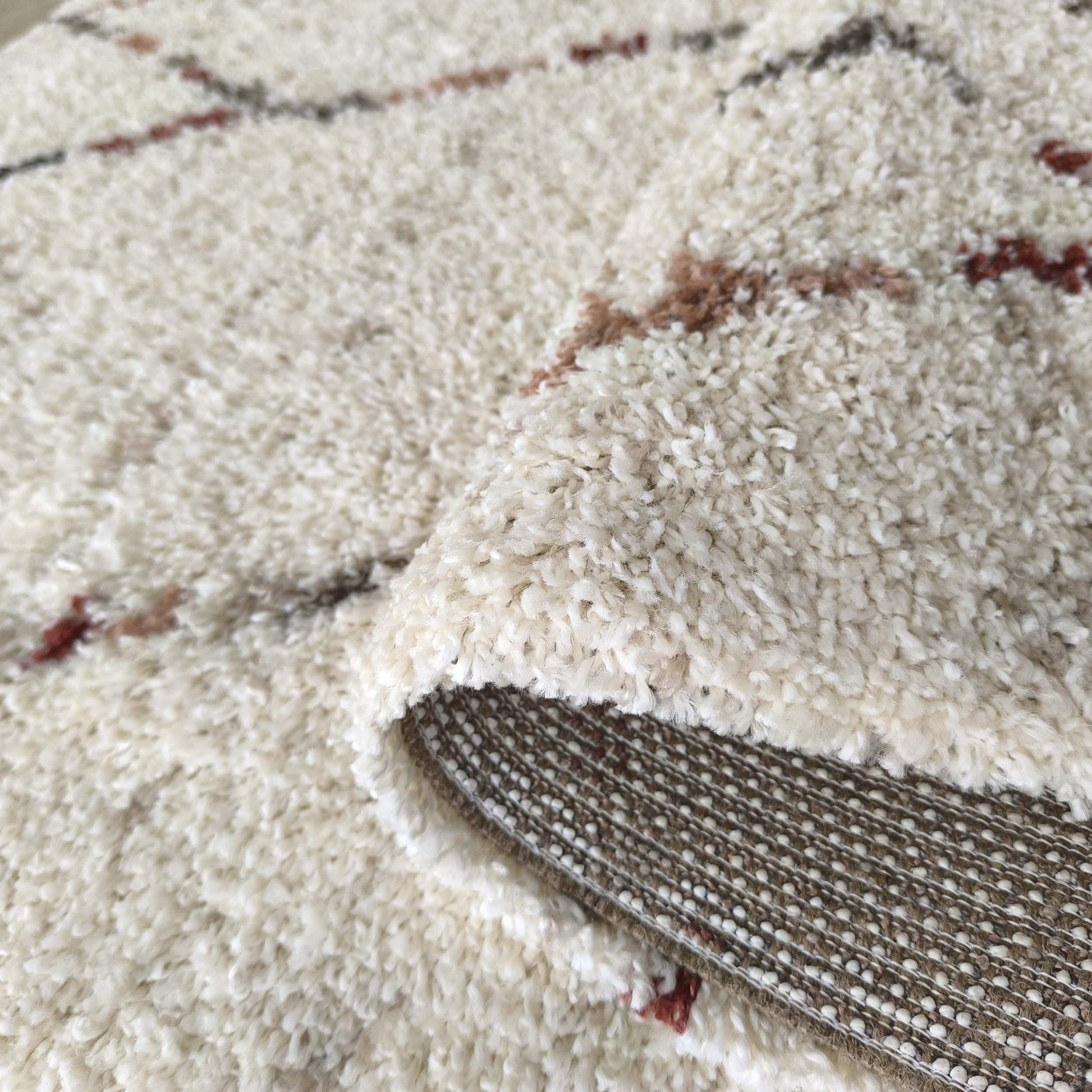 Tapis moelleux blanc cassé aux motifs abstraits marron et rouge. Un coin est replié, révélant son envers tissé