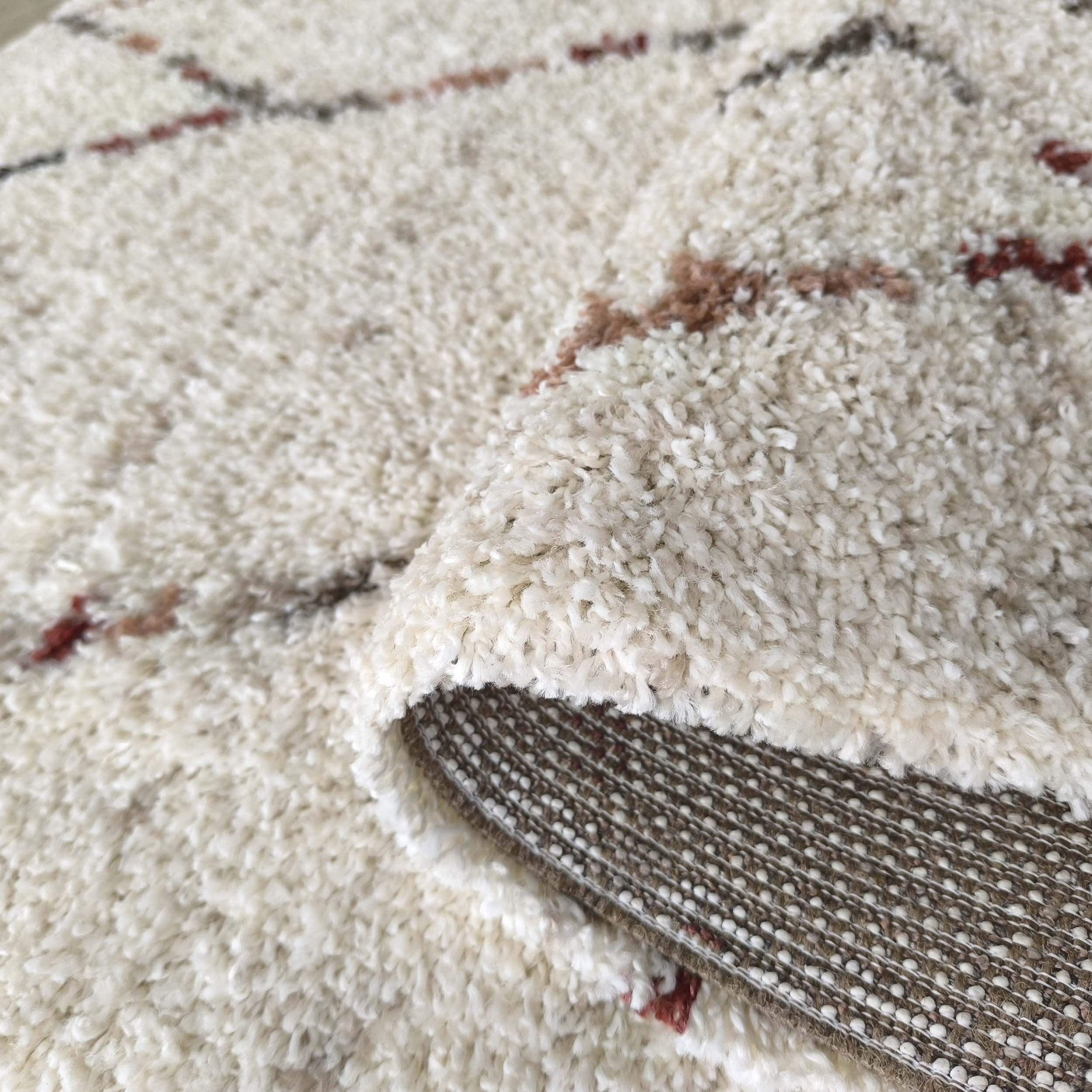 Tapis moelleux blanc cassé aux motifs abstraits marron et rouge. Un coin est replié, révélant son envers tissé