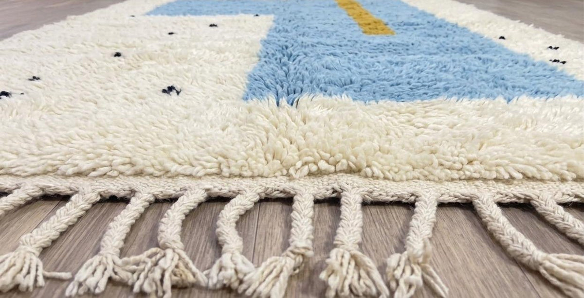 Tapis berbère graphique blanc et bleu ciel Tazmir posé au sol vu de côté, noué main au Maroc