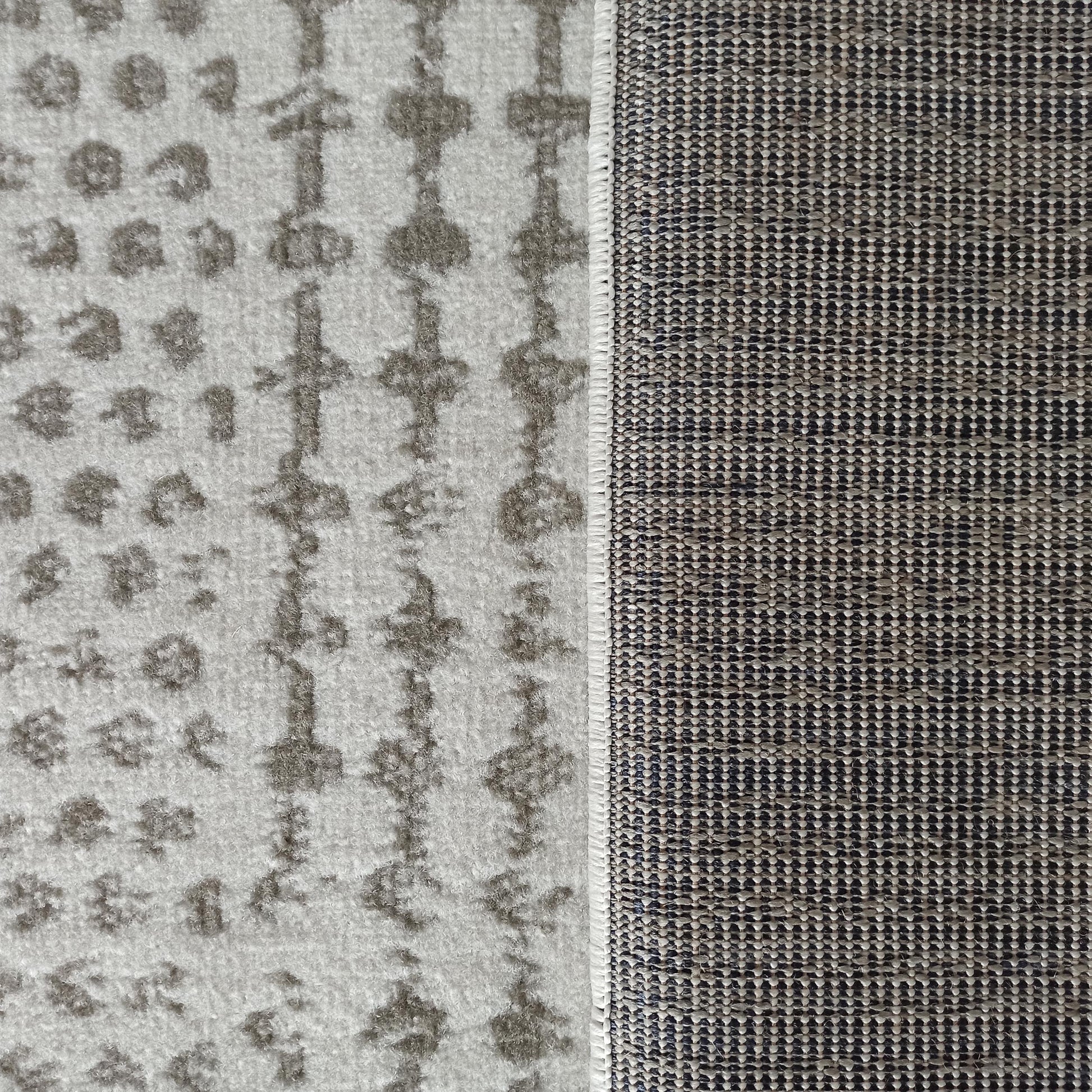 Tapis présentant sa face à motifs abstraits gris clair et foncé, et son envers tissé noir et beige, séparés par une bordure cousue blanche
