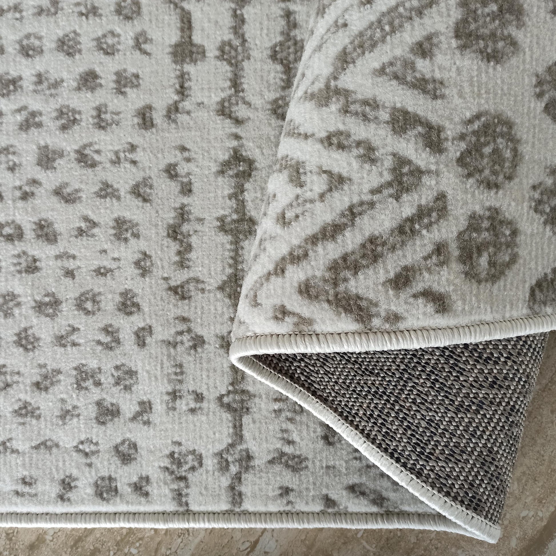 Tapis doux aux motifs abstraits gris foncé sur fond gris clair, avec un coin relevé montrant le dessous tissé