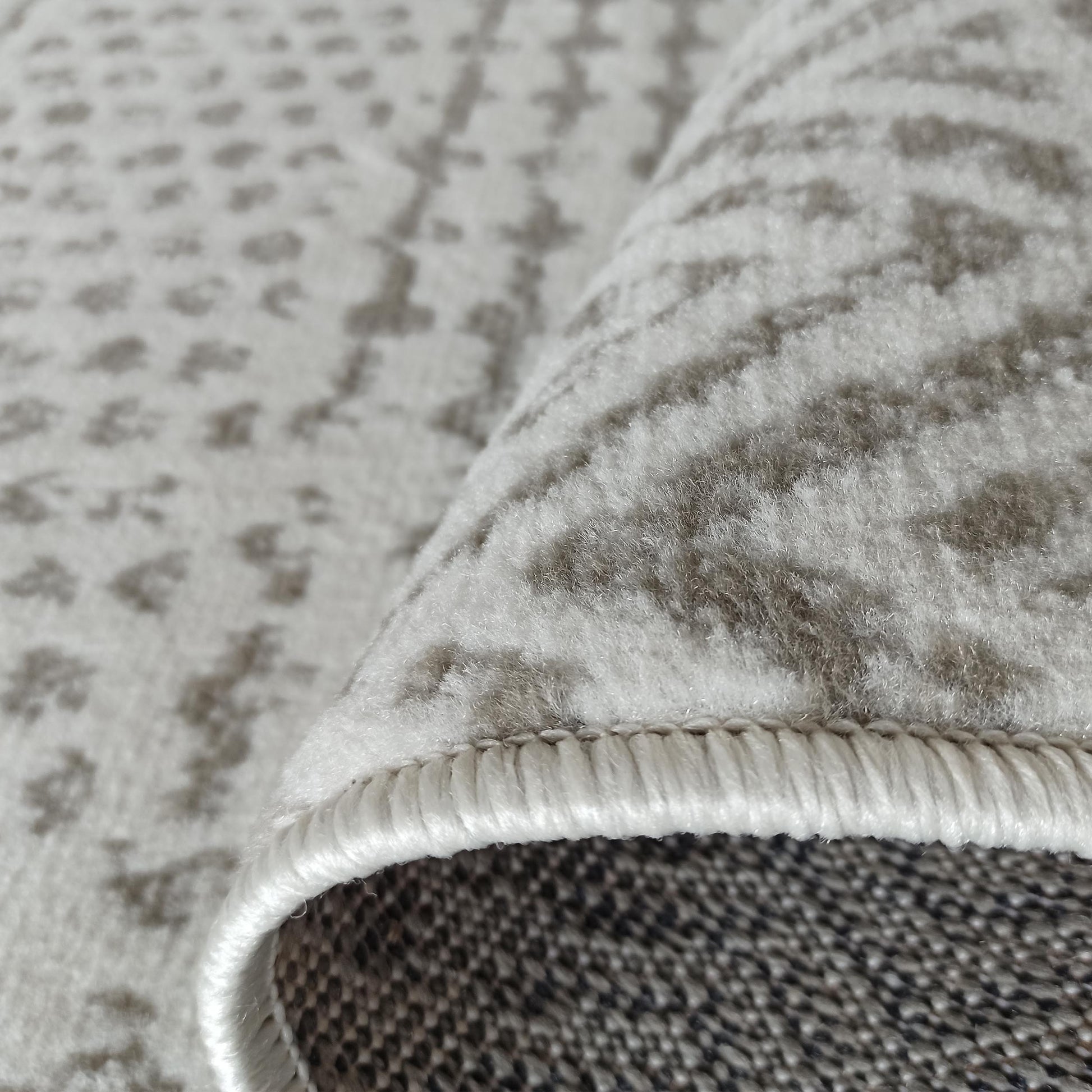 Tapis doux de couleur beige clair orné de motifs gris abstraits, avec un bord roulé montrant l'envers tissé