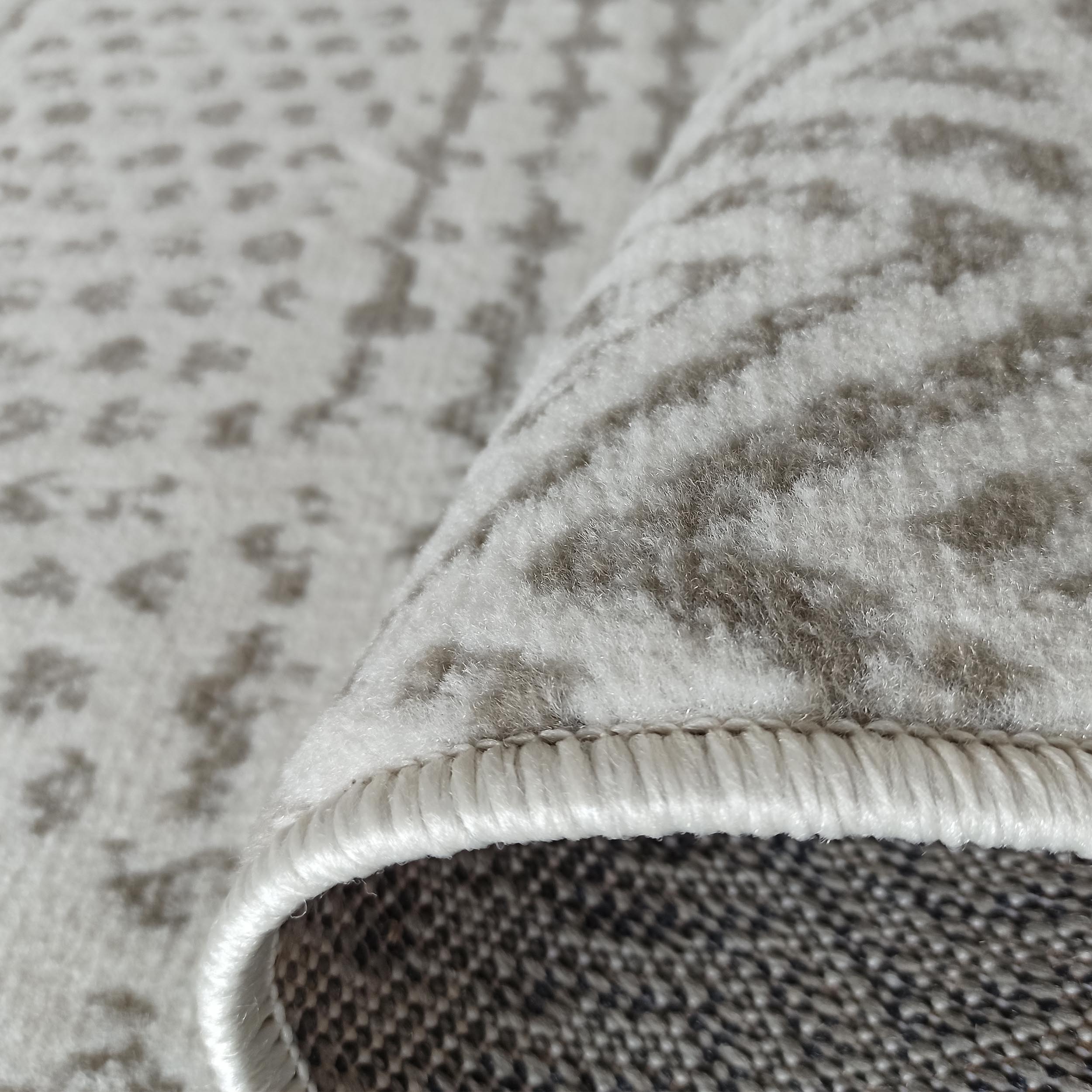 Tapis doux de couleur beige clair orné de motifs gris abstraits, avec un bord roulé montrant l'envers tissé
