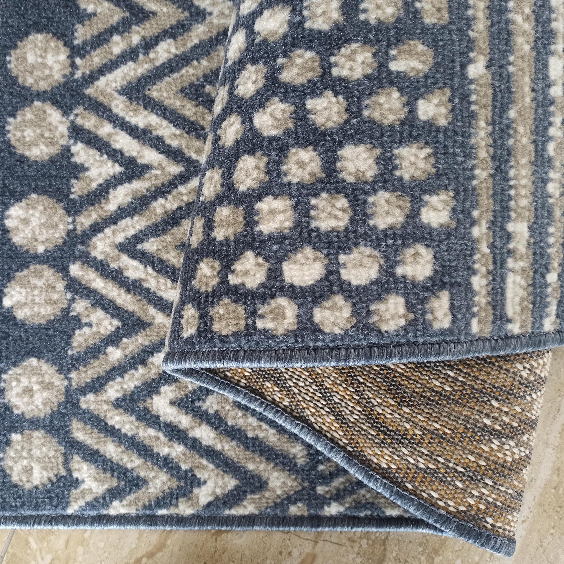Tapis aux motifs géométriques bleus et beiges, zigzags et formes abstraites, partiellement roulé pour révéler son envers tissé