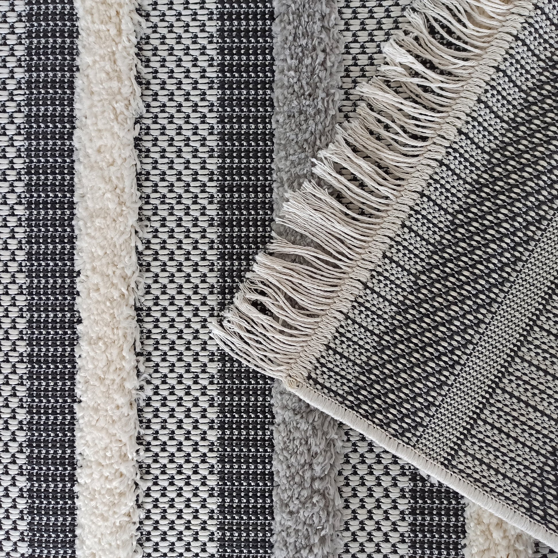 Tapis rayé noir, blanc et gris, composé de bandes à tissage texturé et de bandes à poils longs. Des franges claires sont présentes