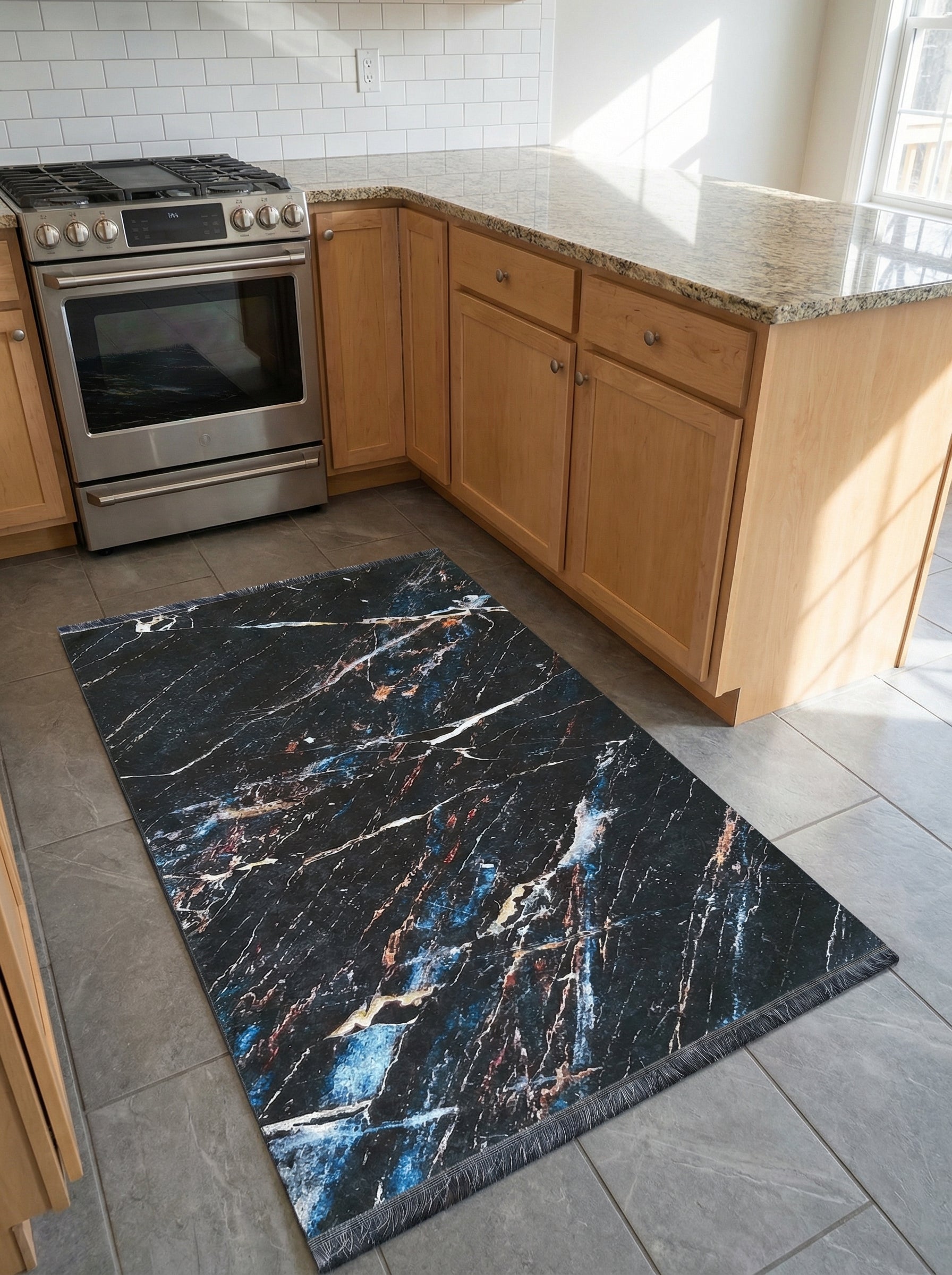 Tapis rectangulaire noir à motifs marbrés blancs, bleus et ocres avec franges, posé sur un sol carrelé dans une cuisine