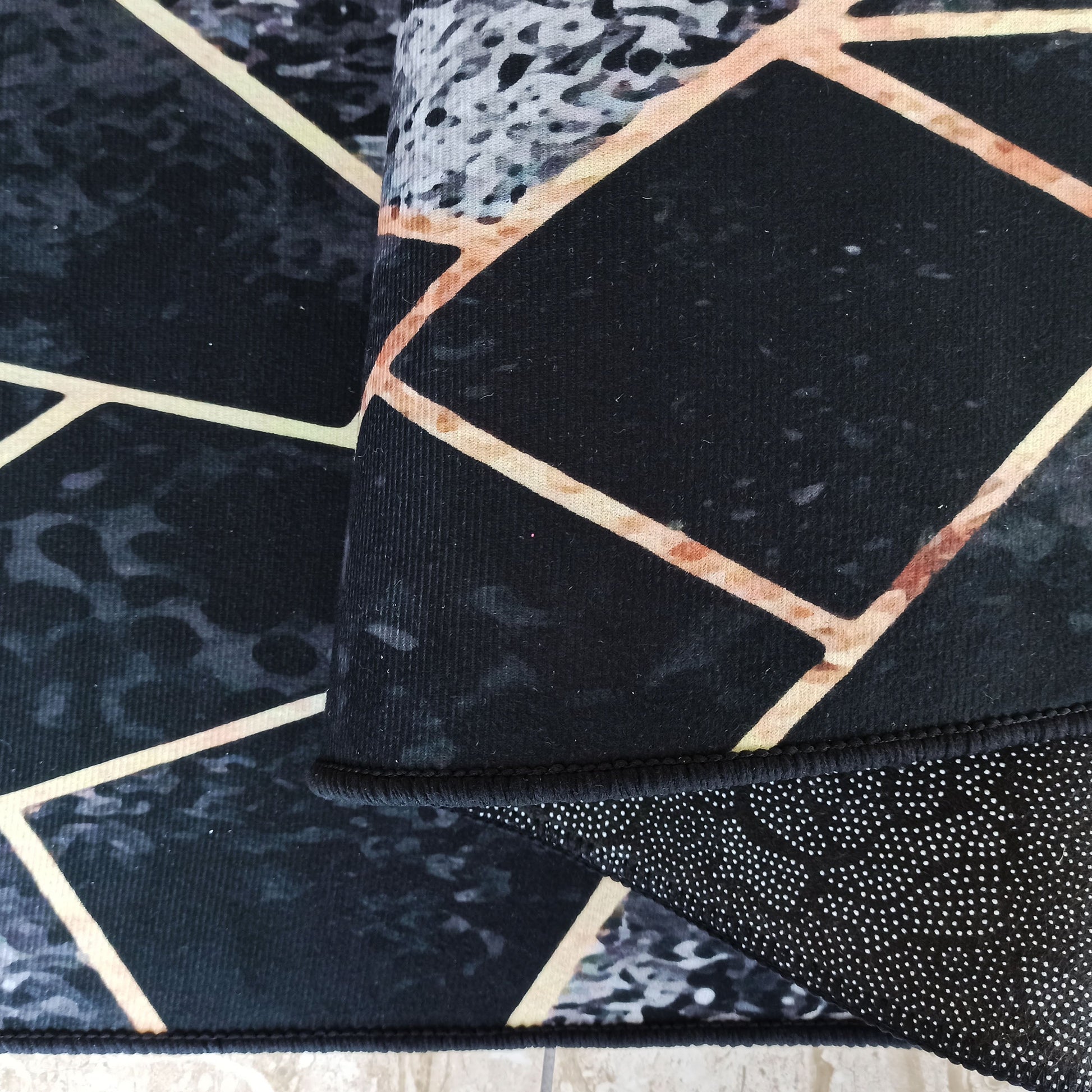 Tapis noir et gris aux motifs géométriques cuivrés et textures marbrées. Le bord et le dessous antidérapant sont apparents