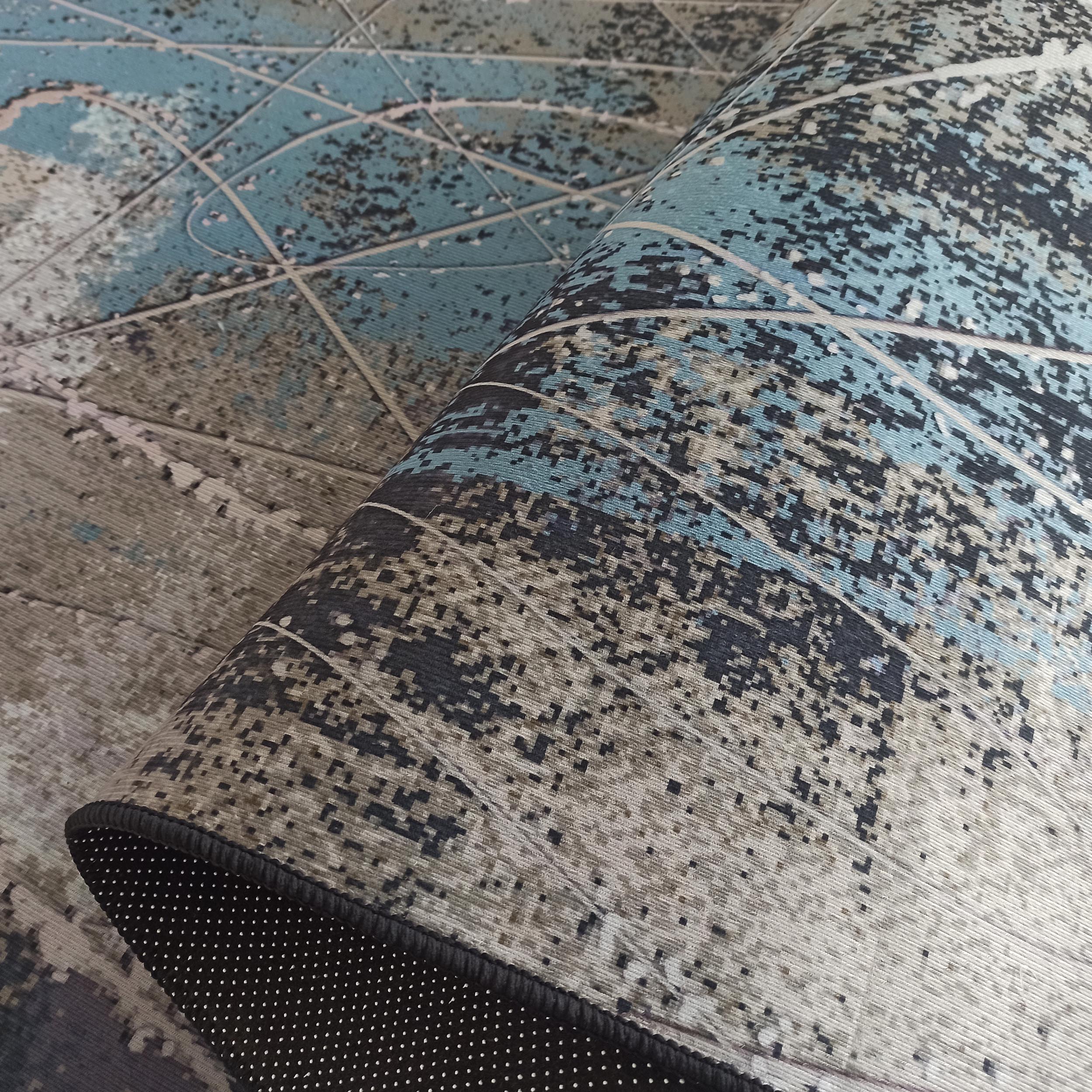 Tapis partiellement roulé aux motifs abstraits bleus, gris et noirs révélant son envers antidérapant noir à pois blancs