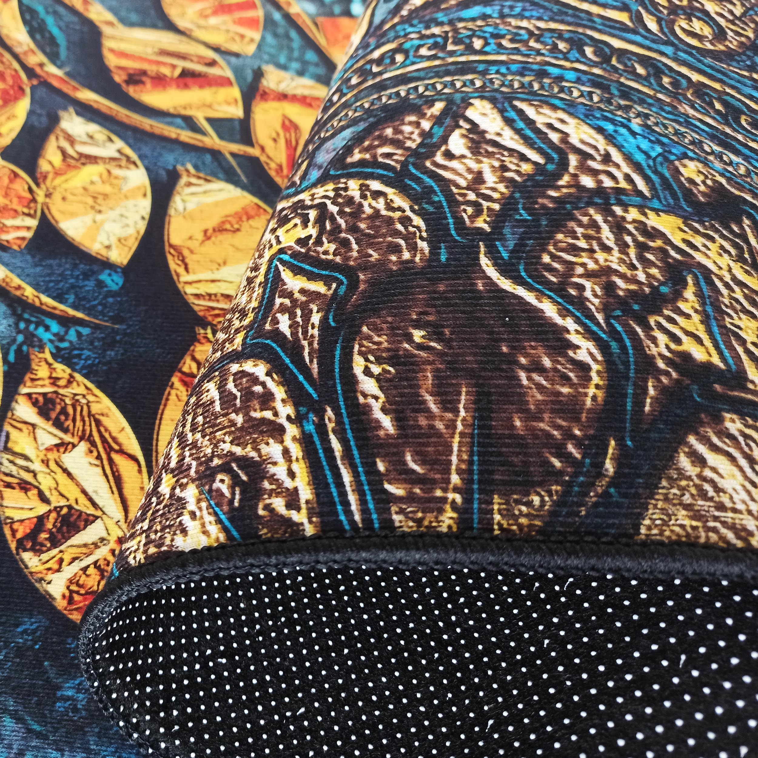 Tapis aux motifs abstraits bleu, jaune et marron, partiellement enroulé. On aperçoit son envers noir à pois blancs et un autre tapis aux feuilles dorées sur fond bleu