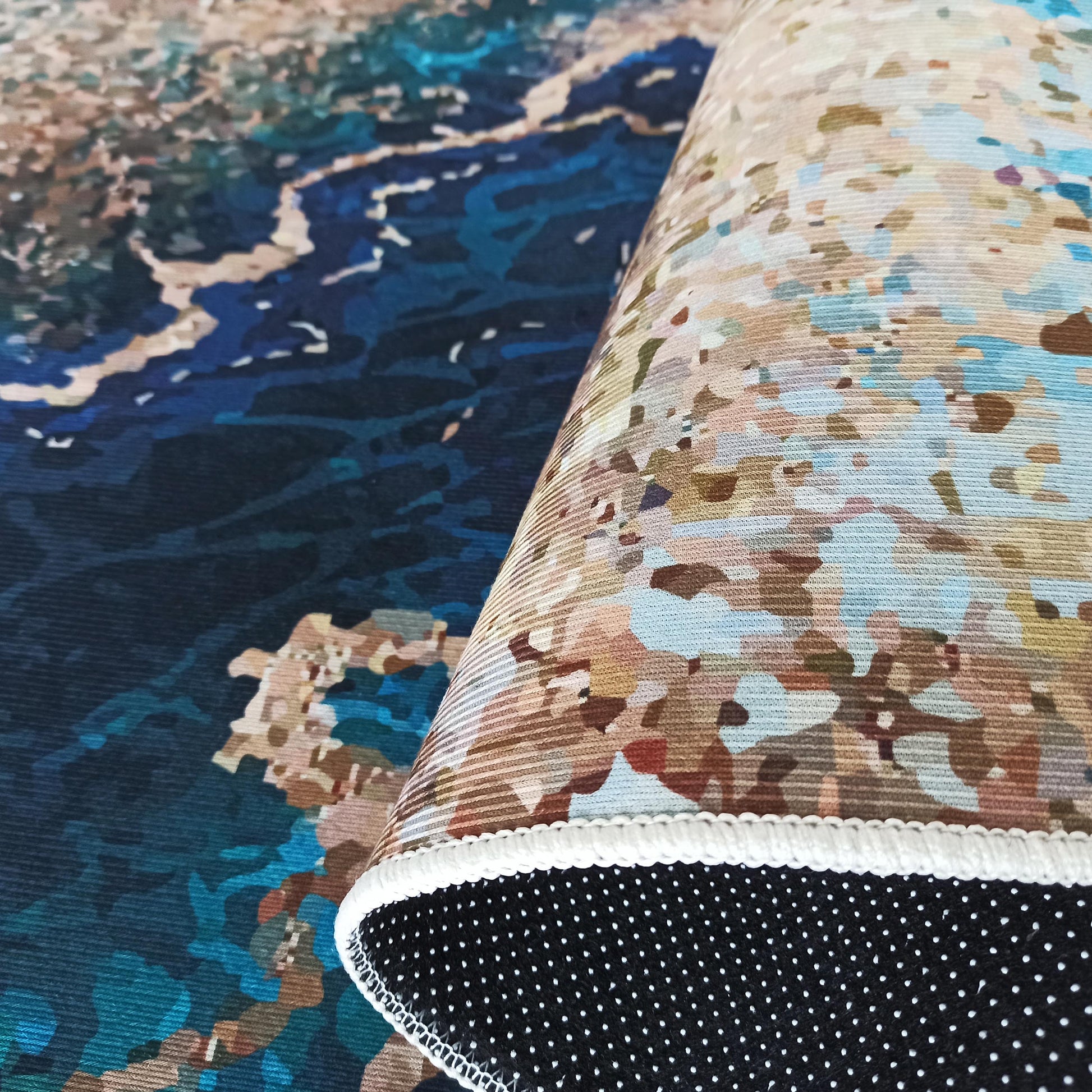 Tapis partiellement enroulé, à motif abstrait de paysages aquatiques et terrestres dans des tons bleus, beiges et marron, avec un dessous antidérapant noir à pois blancs et une bordure festonnée