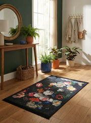 Tapis rectangulaire noir orné de grands motifs floraux multicolores vifs, posé sur un parquet clair dans une entrée lumineuse