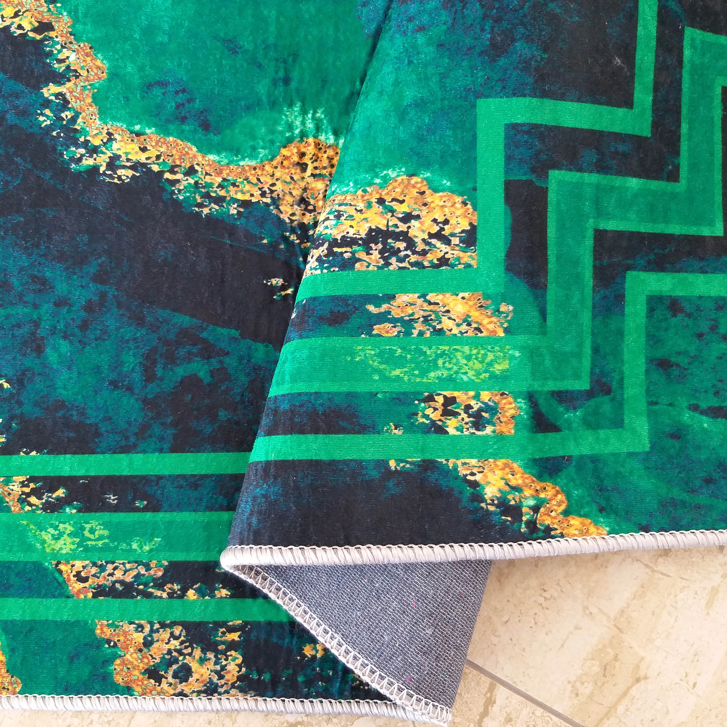 Tapis doux vert émeraude et bleu nuit aux motifs abstraits dorés. Des bandes géométriques vert clair le traversent, son envers gris est visible