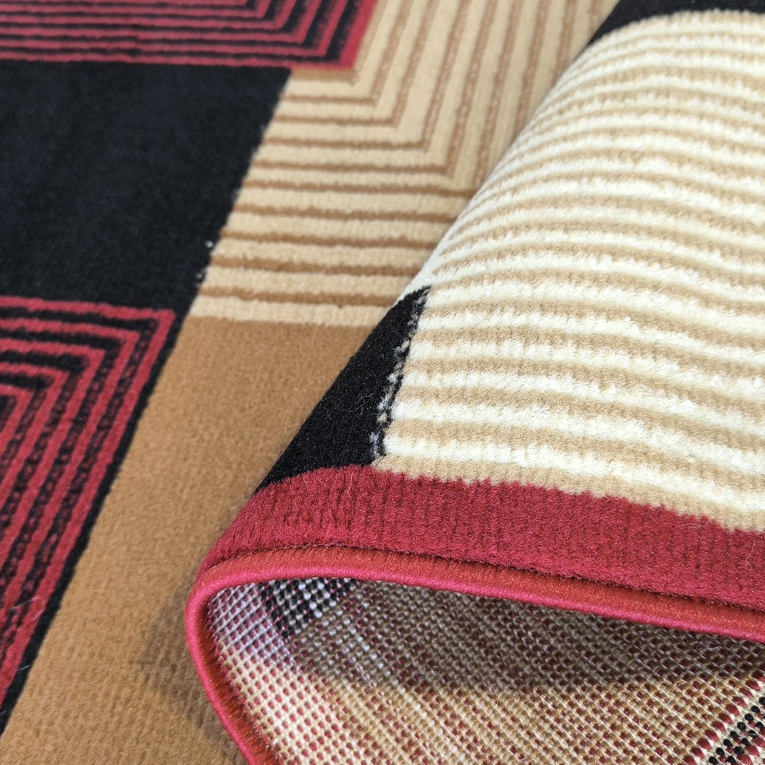 Tapis avec des motifs géométriques rouges, noirs et beiges. Une section roulée révèle des rayures claires et l'envers tissé