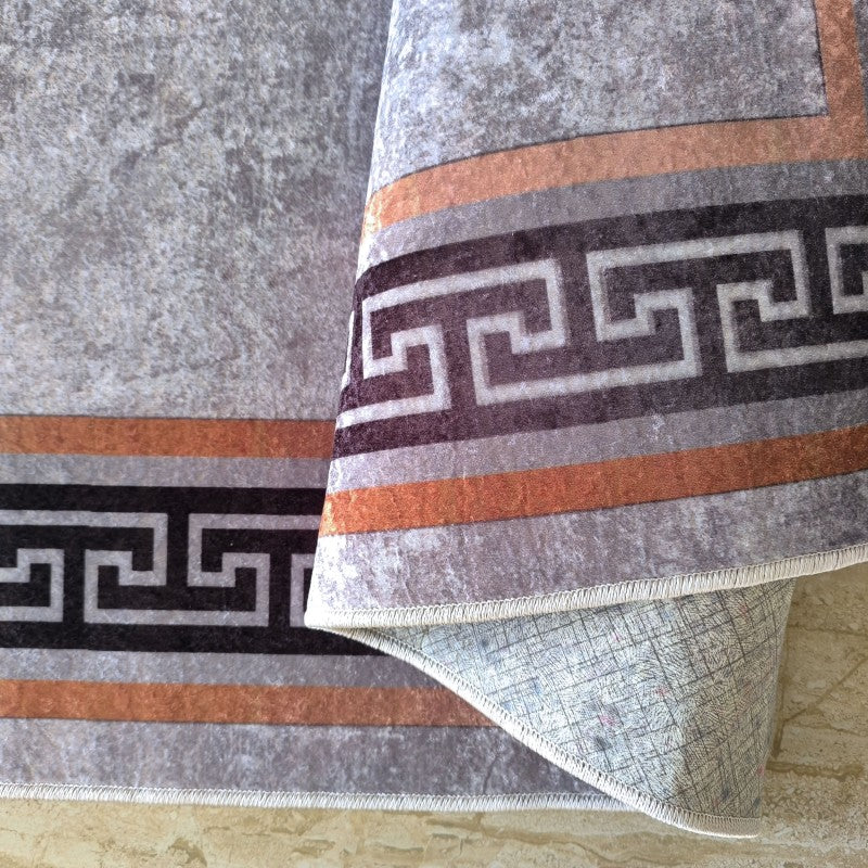 Tapis gris moucheté à bordure géométrique marron foncé et orange rouille, dont un coin est plié, révélant le dessous clair et texturé