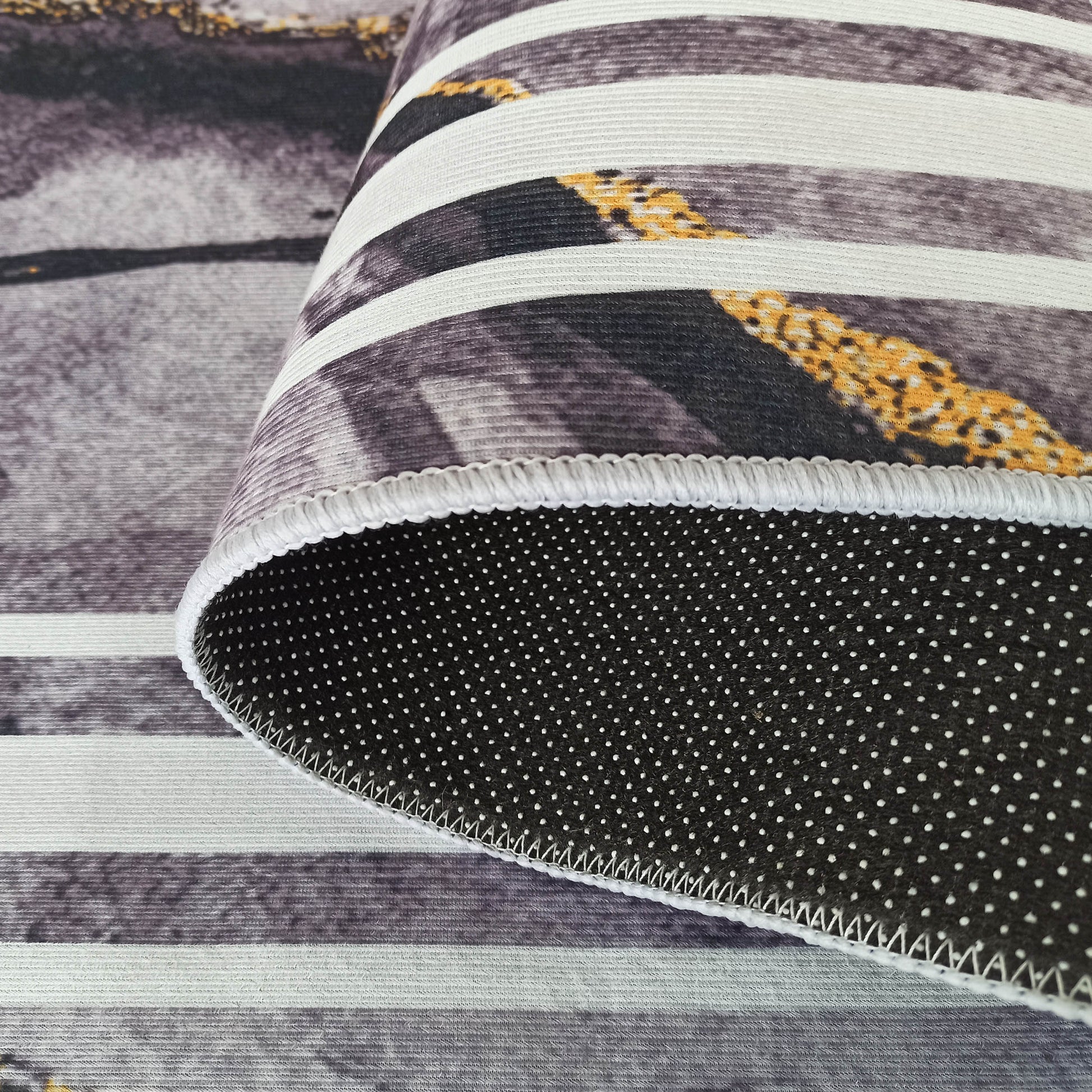Tapis doux à rayures horizontales grises, blanches et tachetées de jaune. L'envers est noir à pois antidérapants blancs