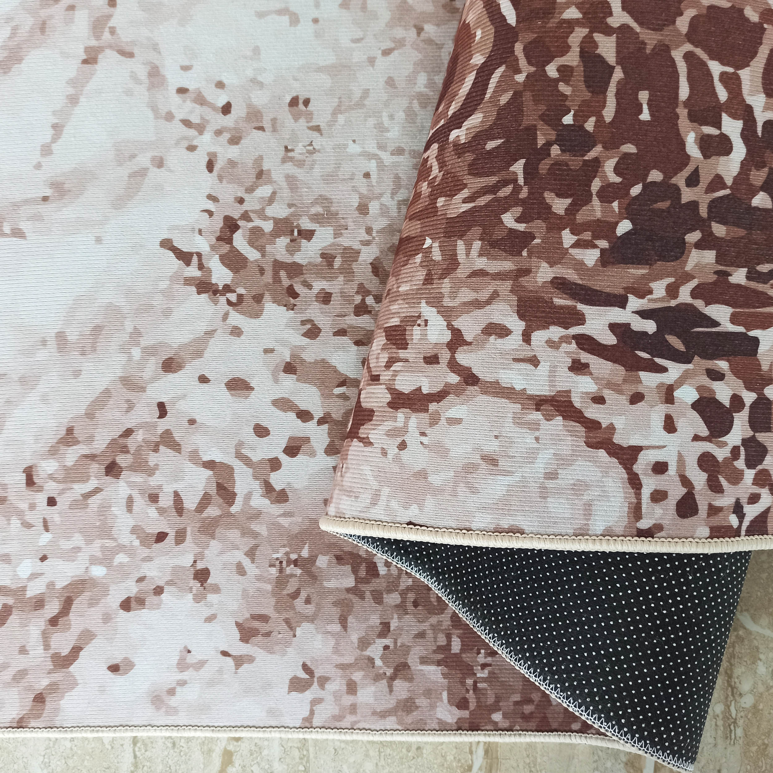 Tapis partiellement roulé à motif abstrait marron et crème, montrant sa face arrière antidérapante noire à pois blancs