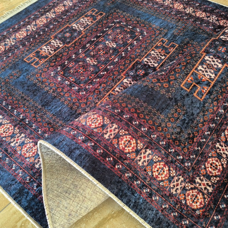 Tapis traditionnel bleu marine orné de motifs géométriques rouges, oranges et crème, avec des franges et un coin soulevé révélant le dos clair