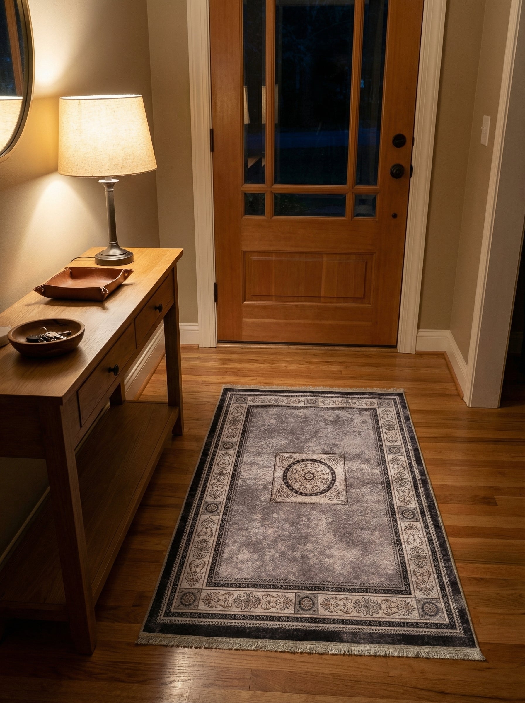 Tapis rectangulaire gris clair, noir et beige, avec un médaillon central et des bordures ornementales, sur parquet en bois dans une entrée